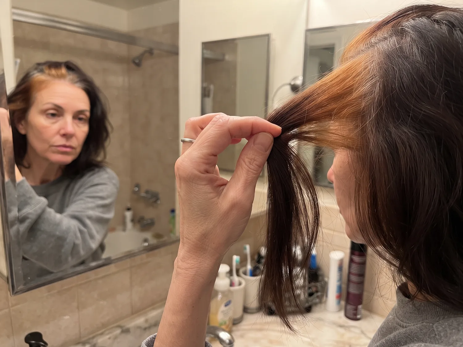 A woman's hand holding a lock of graying hair in bathroom light, the roots tinted a warm copper while the lengths are dark brown